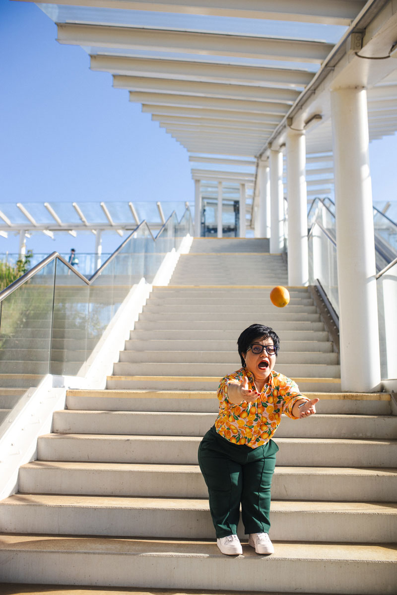 Justyna Cuże łapie w powietrzu pomarańczę na schodach w słońcu.