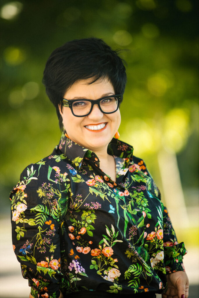 Portret Justyny Cuże w koszuli w kwiaty na tle zieleni drzew.