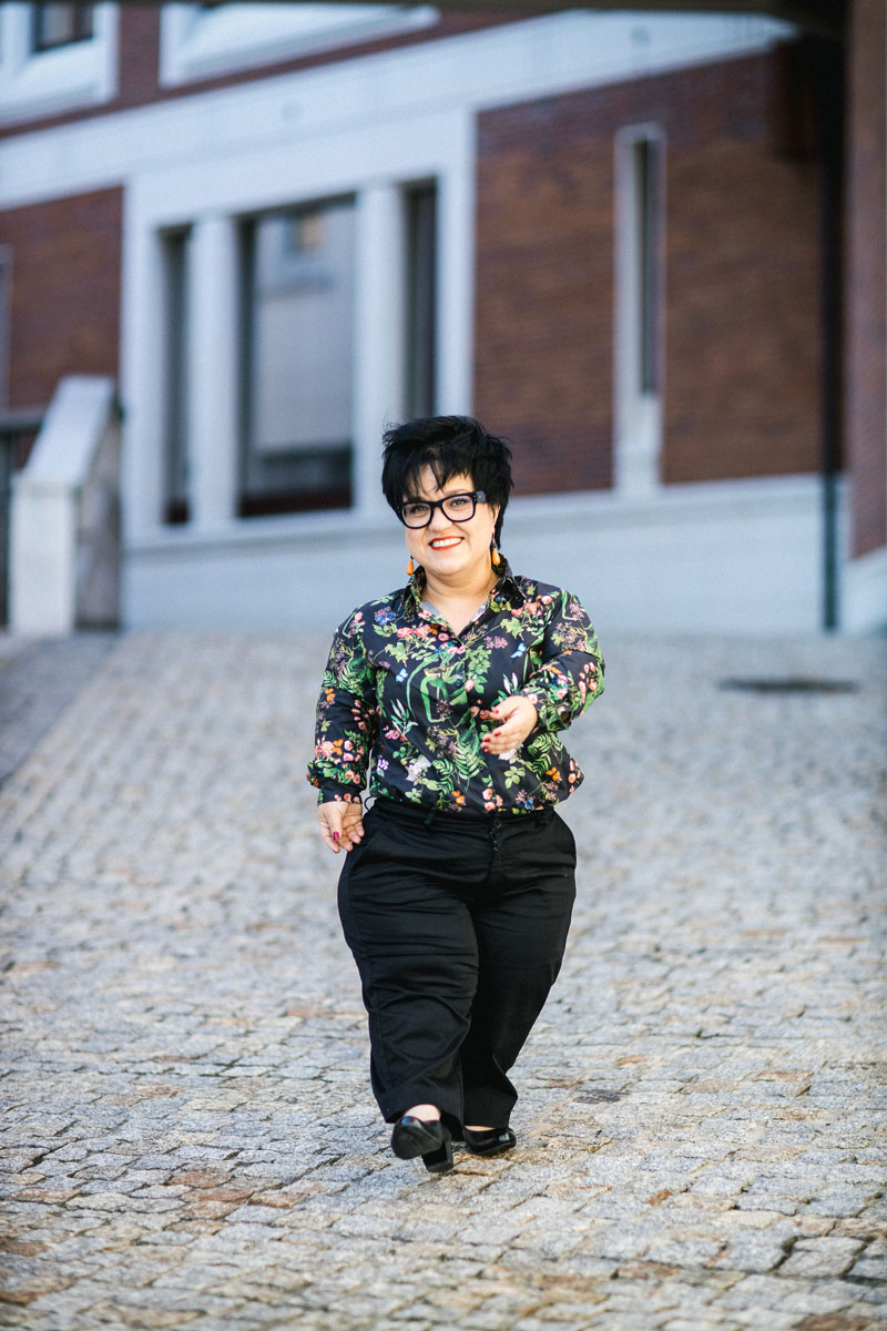 Justyna Cuże idzie po brukowanej uliczce, uśmiechnięta, w kolorowej koszuli.