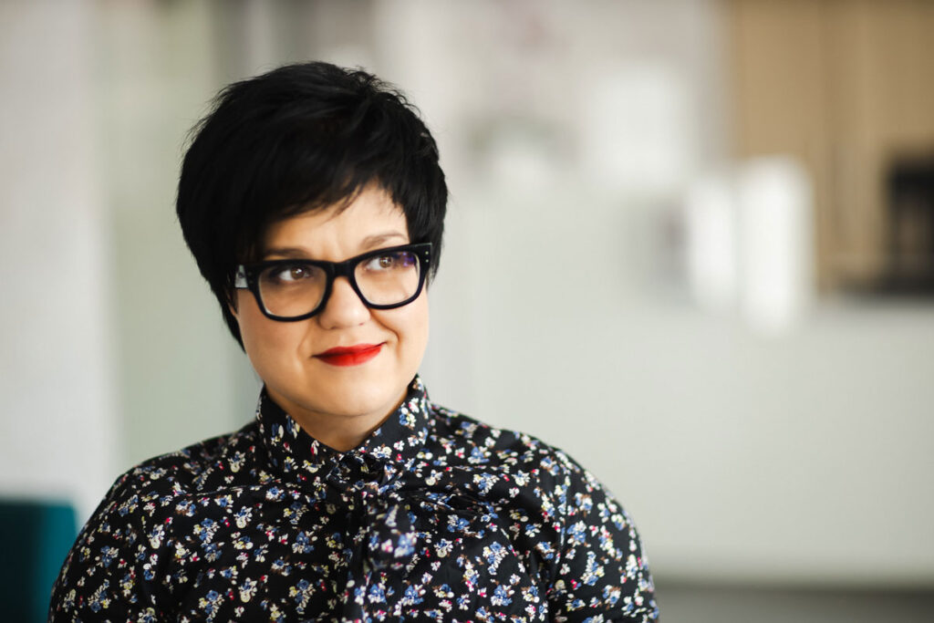 Portret Justyny Cuże w czarnej koszuli w kwiaty, z lekkim uśmiechem.