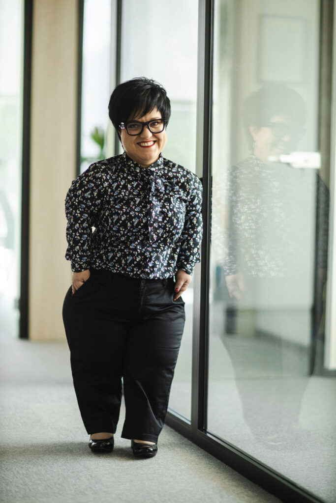 Justyna Cuże stoi w biurowym korytarzu, odbijając się w szybie, z uśmiechem.