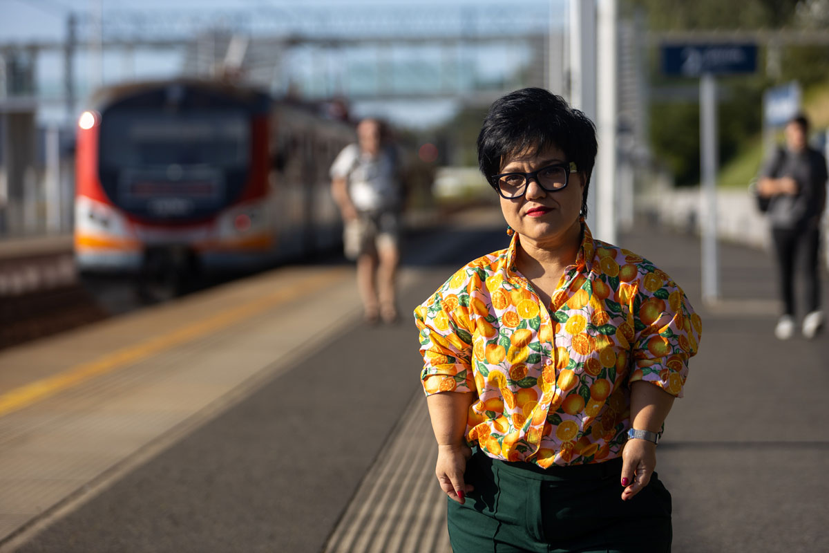 Justyna Cuże stoi na peronie kolejowym w słońcu, w kolorowej koszuli w owoce.