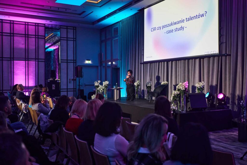 Justyna Cuże stoi na scenie konferencji, prowadząc prezentację „CSR czy poszukiwanie talentów? – case study”.