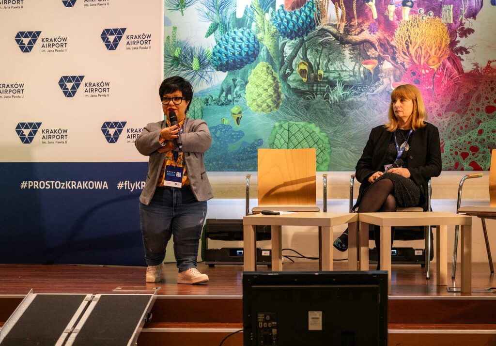 Justyna Cuże stoi na scenie z mikrofonem, obok siedzi kobieta, w tle baner Kraków Airport i kolorowa grafika.