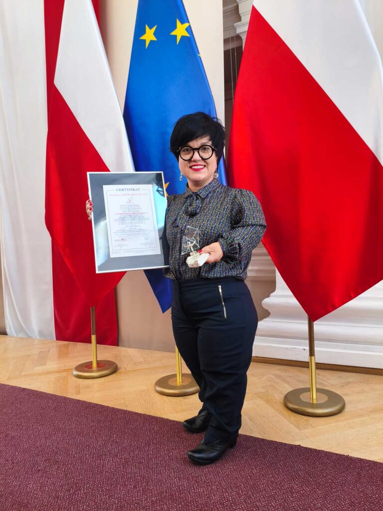 Justyna Cuże pozuje z certyfikatem i statuetką Lodołamacza na tle flag Polski i Unii Europejskiej.