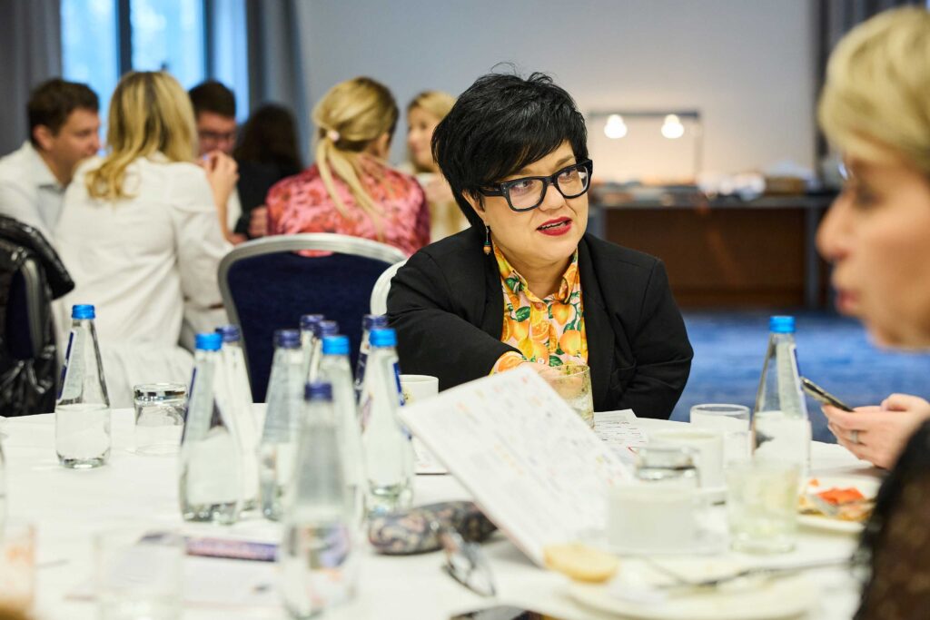 Justyna Cuże rozmawia przy stole podczas spotkania, na którym uczestnicy siedzą w grupach i prowadzą dyskusje.