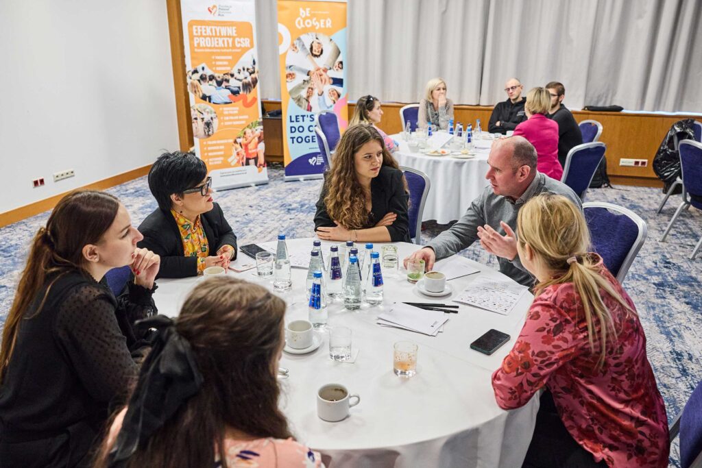 Grupa sześciu osób, w tym Justyna Cuże, dyskutuje przy okrągłym stole na warsztatach dotyczących projektów CSR.