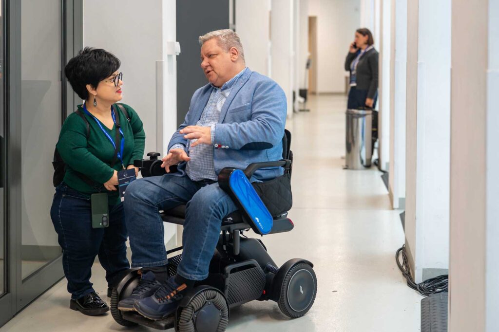 Justyna Cuże rozmawia z mężczyzną na wózku elektrycznym w jasnym korytarzu biurowym.
