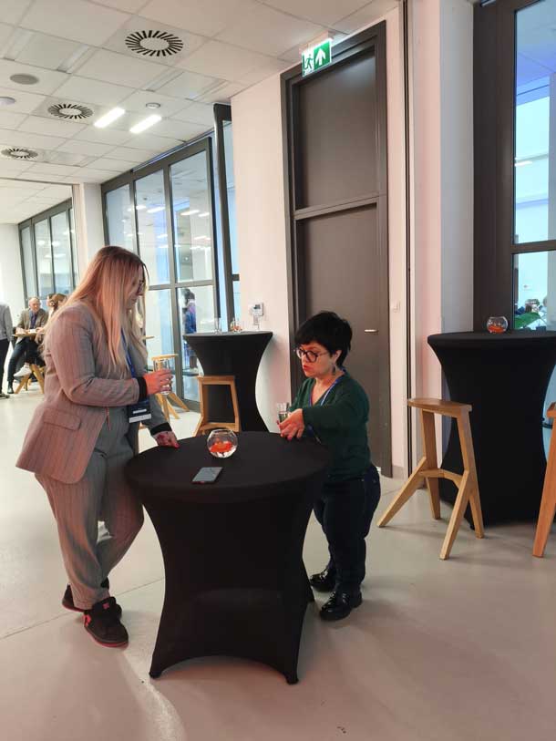 Justyna Cuże stoi przy stoliku i rozmawia z kobietą w jasnym garniturze w przestrzeni konferencyjnej.