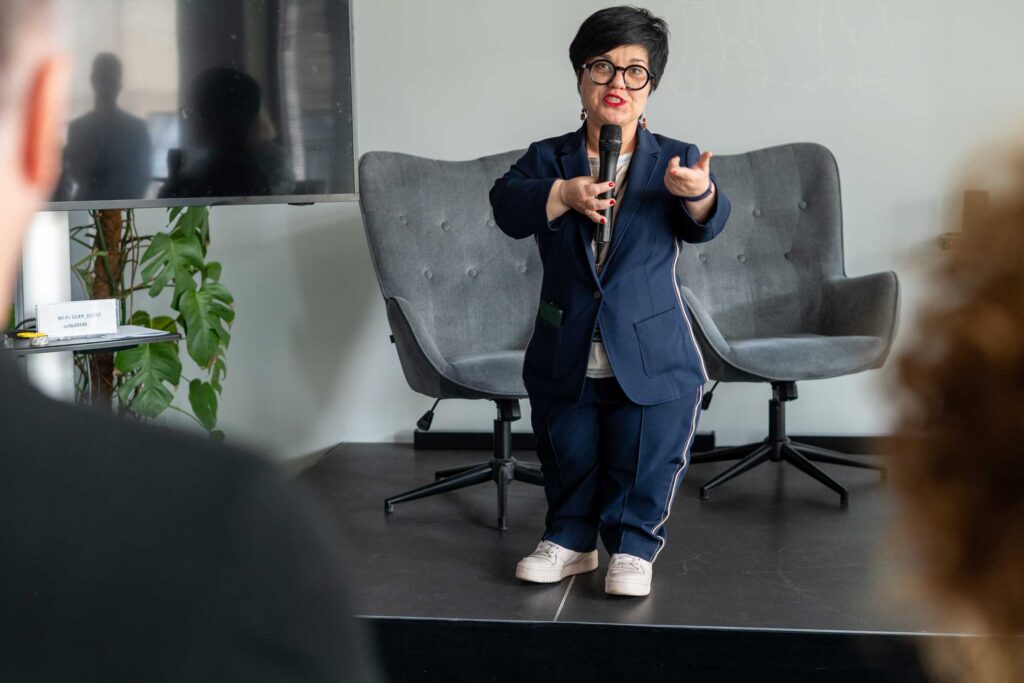 Justyna Cuże prowadzi wystąpienie, stojąc na scenie z mikrofonem w dłoni.